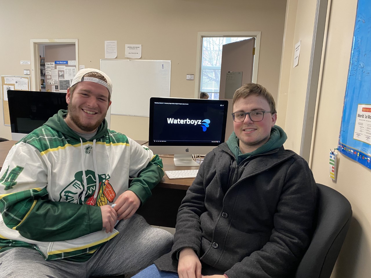 grant and justin pose in front of their computer in the journalism classroom