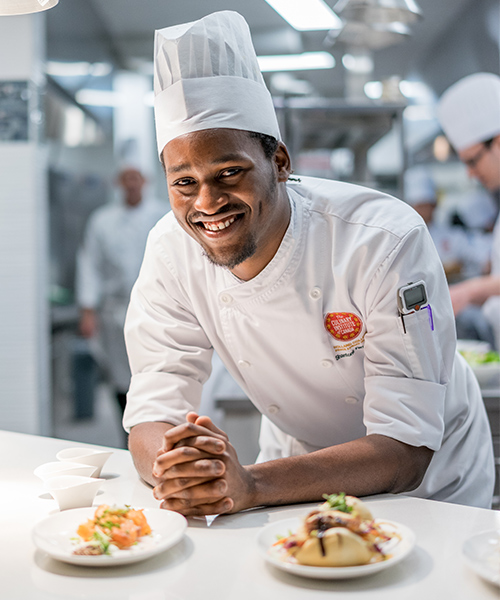 black male student chef leaning forward on the countertop alongside plated meals 